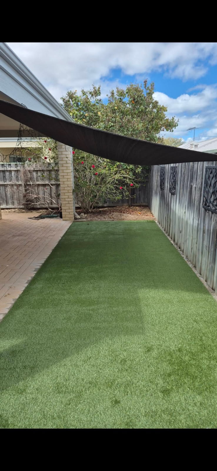 A strip of artificial grass laid alongside a house with a shade sail overhead